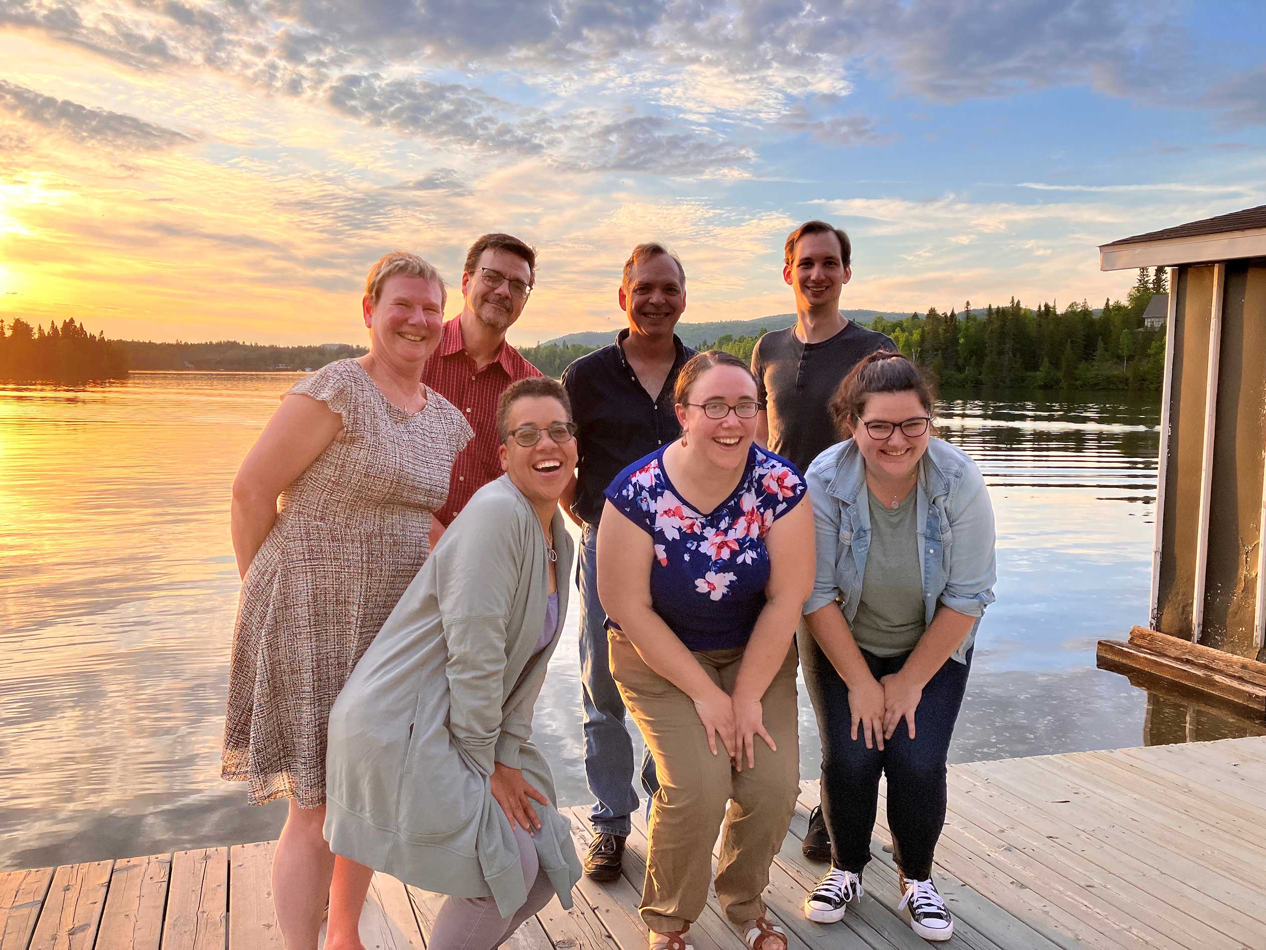 Seven alumni pose on the dock of a lake, with the setting sun in the background.