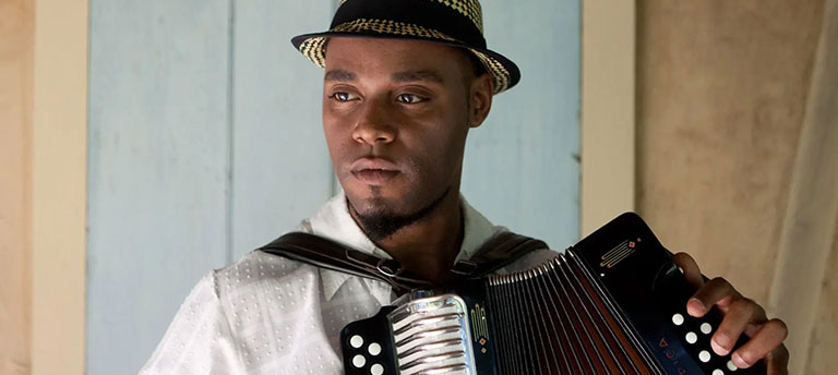 A headshot of Cedric Watson, who wears a white shirt and holds an accordion. 