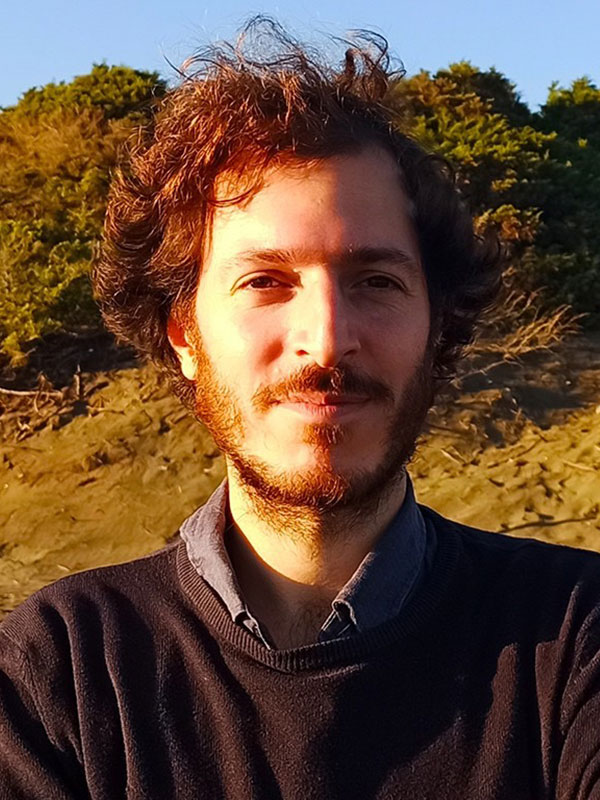 A headshot of Filippo Petricca, who wears a brown sweater and poses outside.