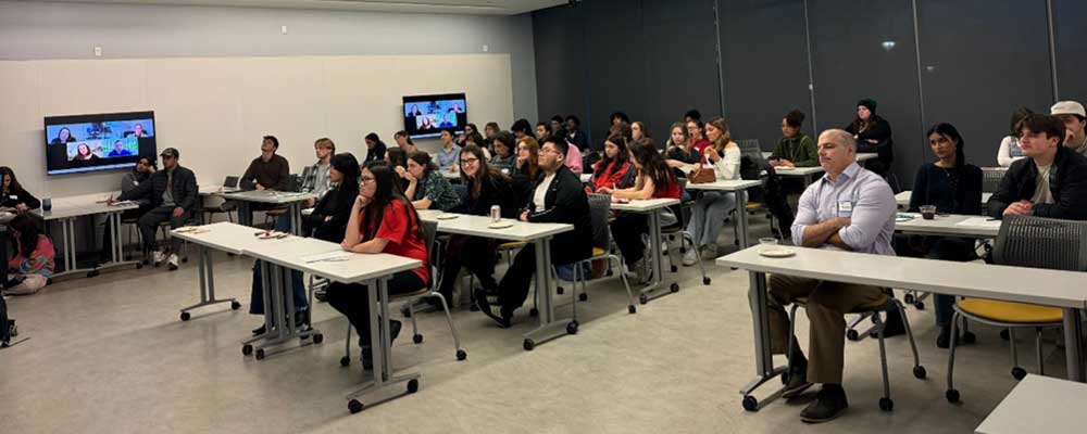 A classroom full of students listens attentively to a panel discussion, with two screens displaying remote speakers.