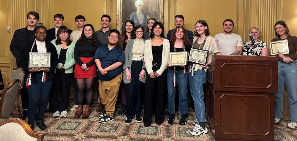 A group of students and faculty pose for a photo during an awards ceremony, with some holding certificates and cords.