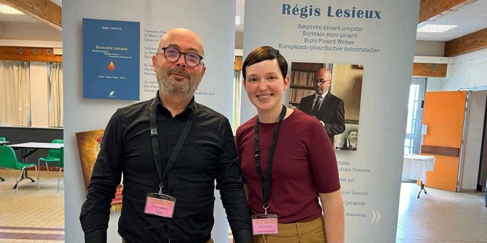 Two people smile and pose in front of a display about Régis Lesieux at a language-focused event.