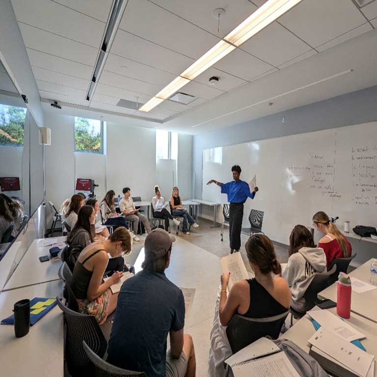 A professor gestures expressively while teaching a classroom of students seated in a semicircle with notebooks.