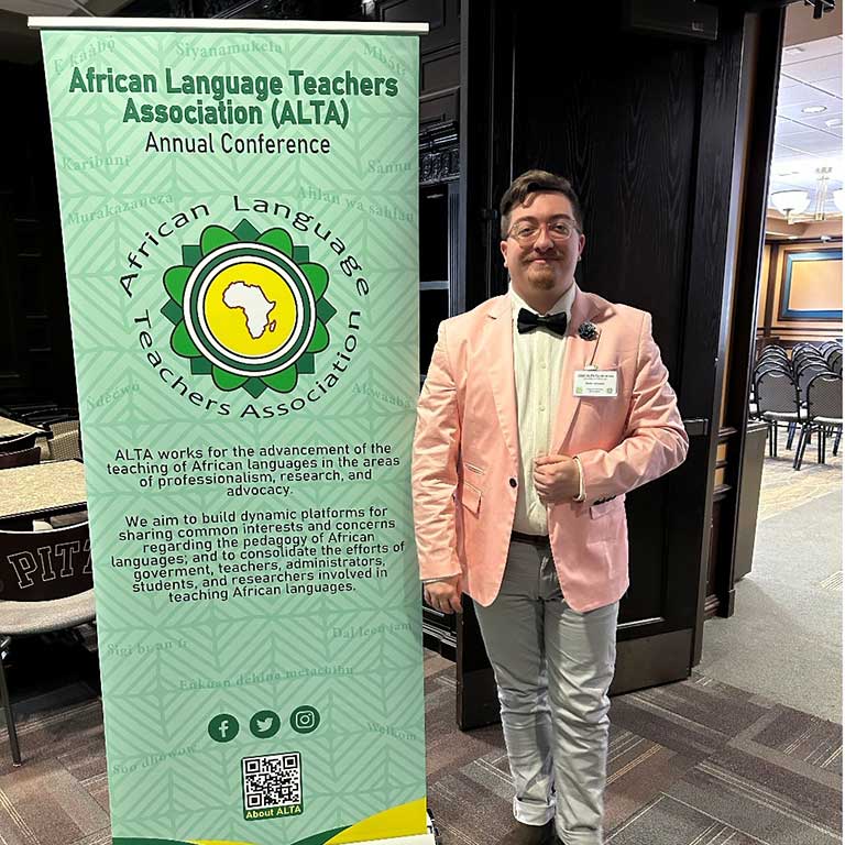 A man in a pink blazer and bow tie stands beside a banner for the African Language Teachers Association Annual Conference.