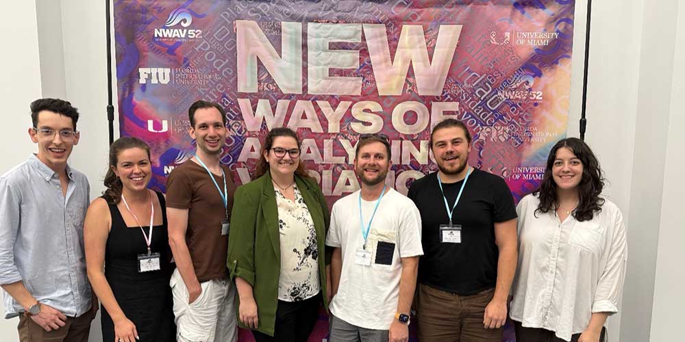 A group of seven conference attendees pose together in front of a colorful banner reading "New Ways of Analyzing Variation."
